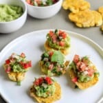 A plate of crispy tostones on the table with bowls of guacamole and tomatoes in the background.