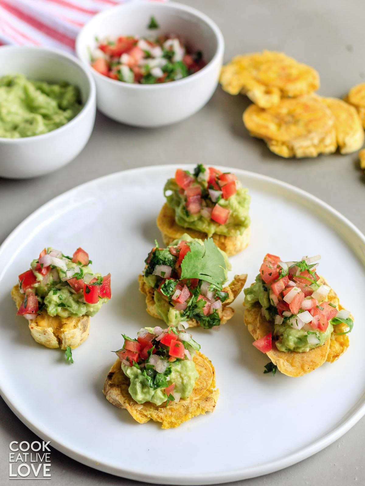A plate of crispy tostones on the table with bowls of guacamole and tomatoes in the background.