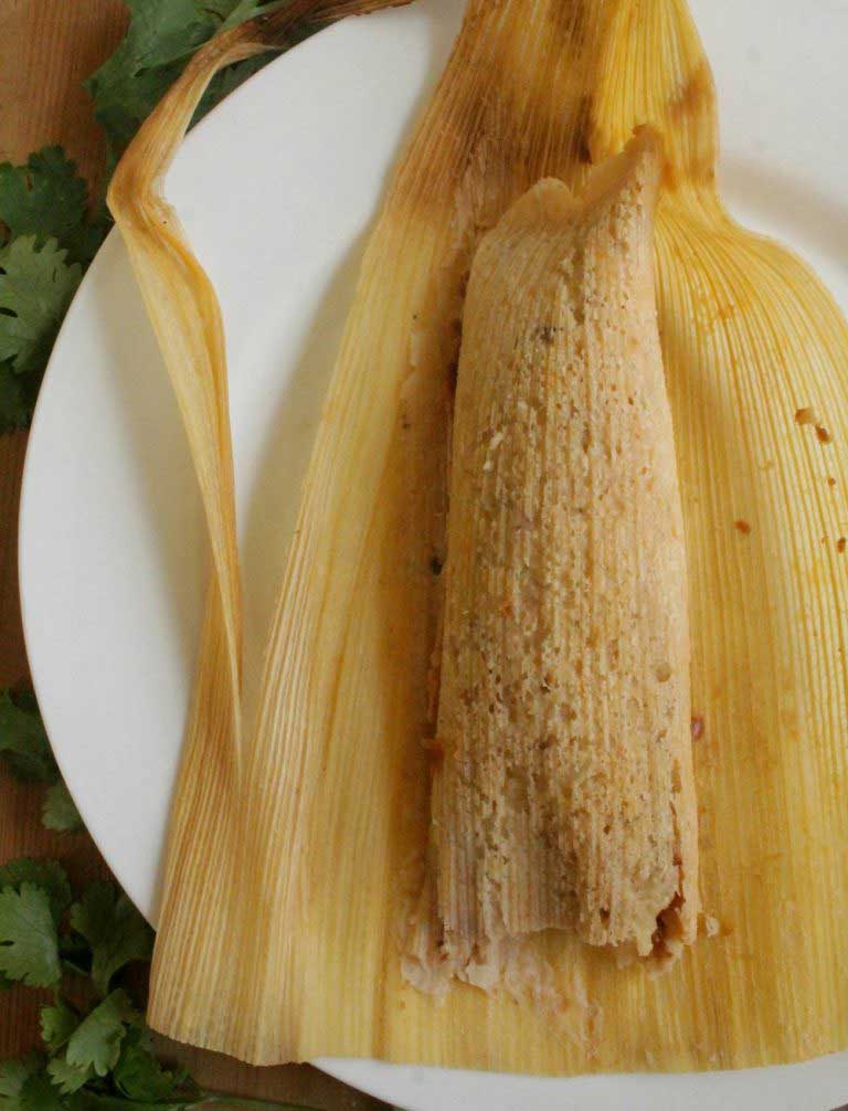 Tamale in corn husk displayed on white plate.
