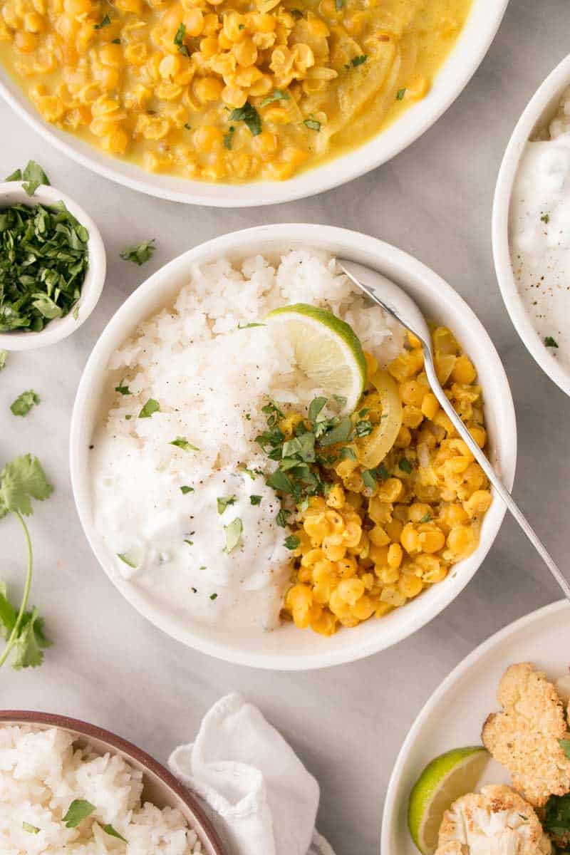 Bowl of yellow dal and white rice displayed on white background. Bowl is garnished with lime and chopped parsley.