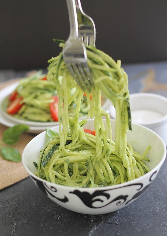 Bowl of zucchini noodles with avocado sauce. Above bowl, two forks are lifting noodles up from bowl.