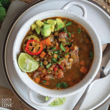 Bowl of Mexican lentil soup on a plate with a spoon topped with avocado, chili slices and lime.