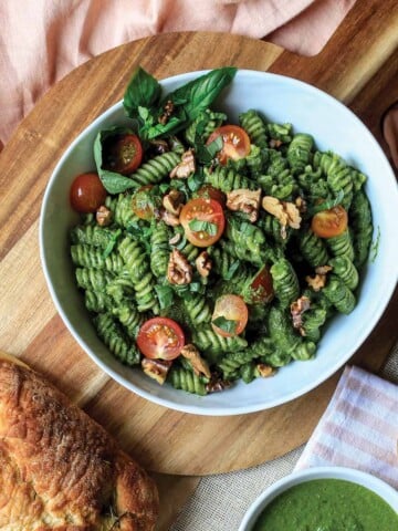 A bowl of spinach pesto pasta on the table with tomatoes and basil.