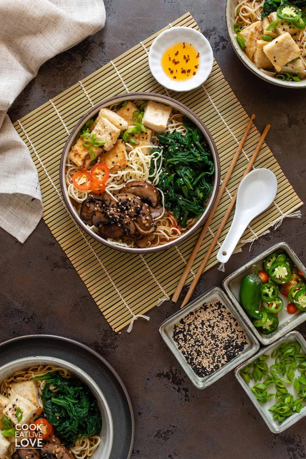 Vegan tofu ramen bowl on the table with toppings, chopsticks and a spoon.