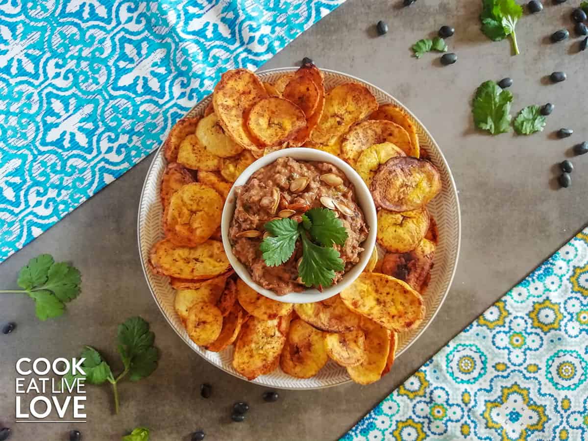 Platter with bowl of black bean hummus surrounded by plantain chips .