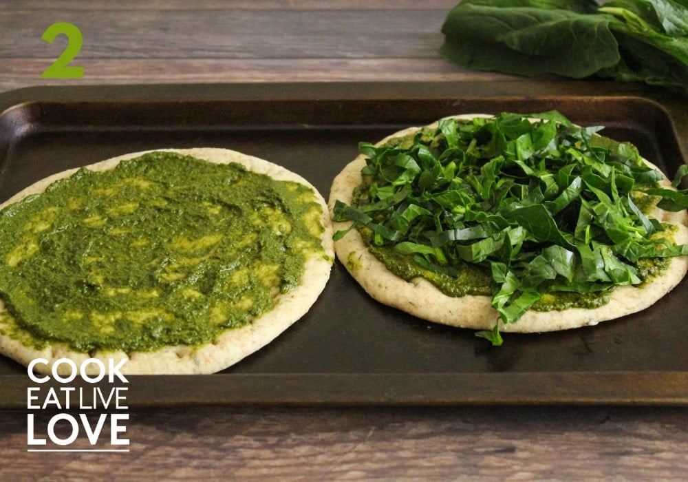 Pizzas on pan, pesto is spread and then spinach is added.