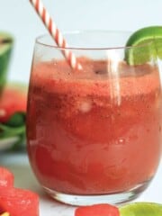 Close up of watermelon slushie in a glass with watermelon slices and pitcher of more slushie behind.