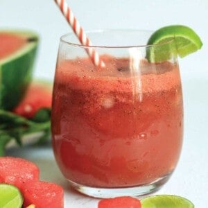 Close up of watermelon slushie in a glass with watermelon slices and pitcher of more slushie behind.