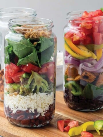 Glass jar with layered grain and vegetaables. Whole tomatoes and spoon on counter in front and piles of fresh vegetables behind.