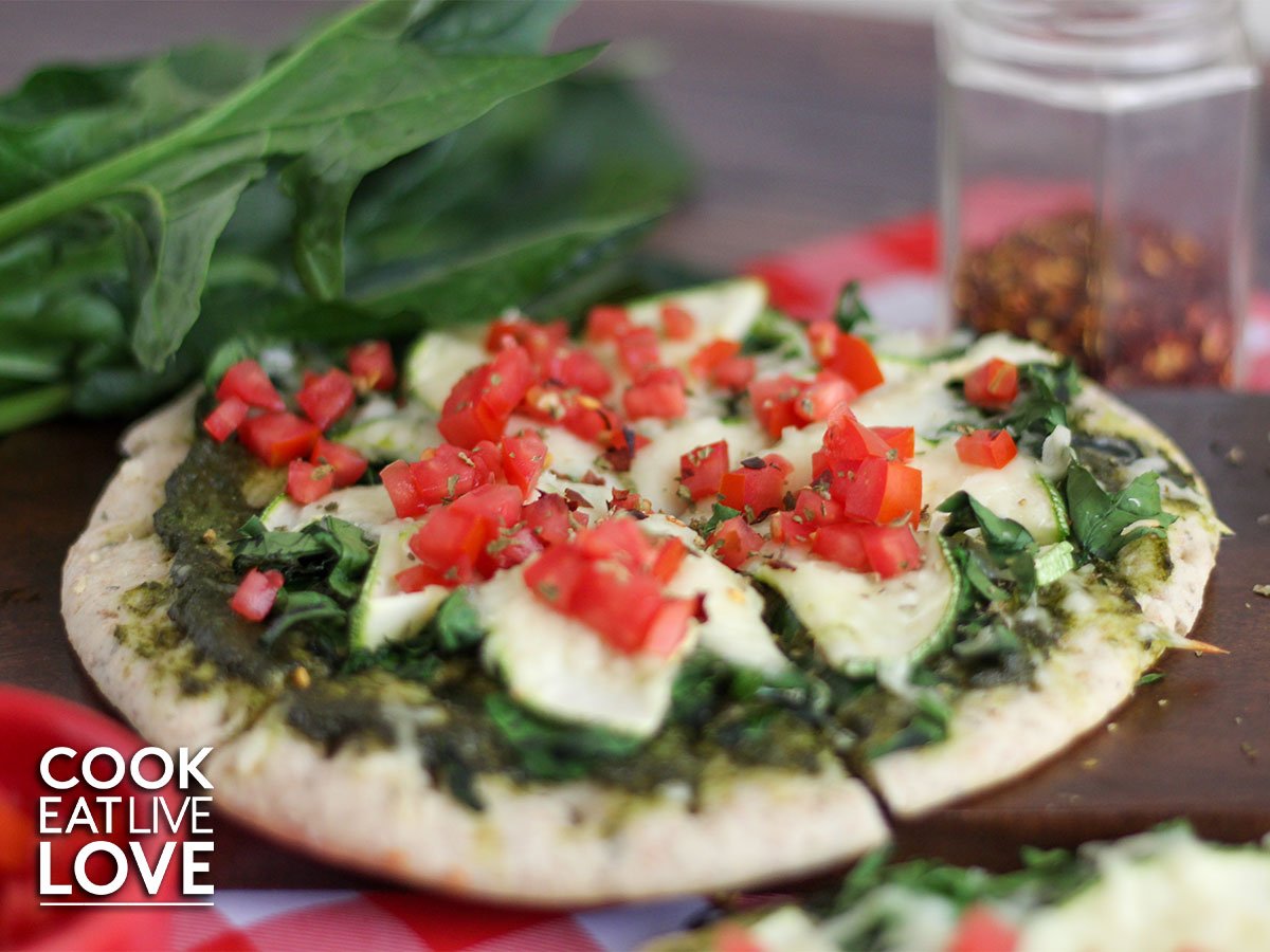 Pesto pita pizza is cooked on cutting board with spinach, crushed red pepper in background 