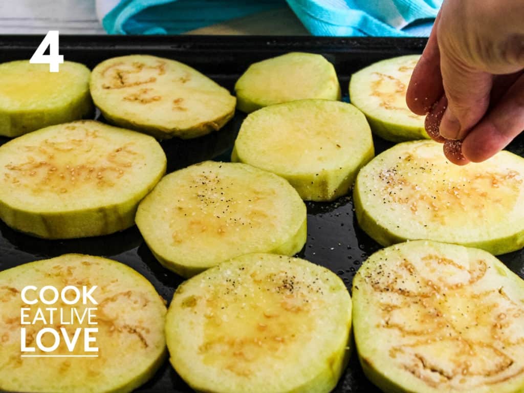 Eggplant topped with olive oil are shown on baking sheet.  A hand is shown sprinkling salt and pepper on the eggplant.