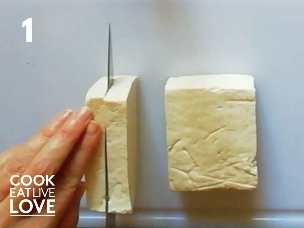 Block of tofu is on a cutting board.  Knife is cutting it into ¼ inch slices.
