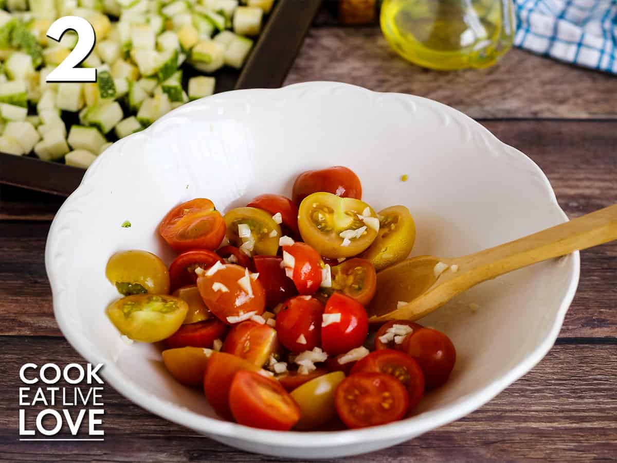 Tomatoes cut in half with olive oil and garlic in a white bowl. With wooden spoon in the bowl and the zucchini on the baking sheet in the top corner.