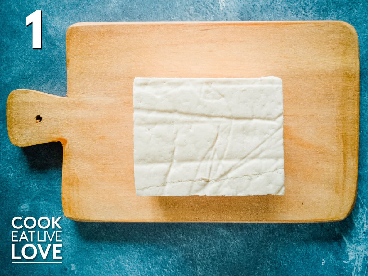 Block of tofu out of container and sitting on wooden cutting board on blue background.