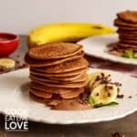 Front view of stack of 10 pancakes on a white plate with sliced bananas, cacao cream and fresh mint.