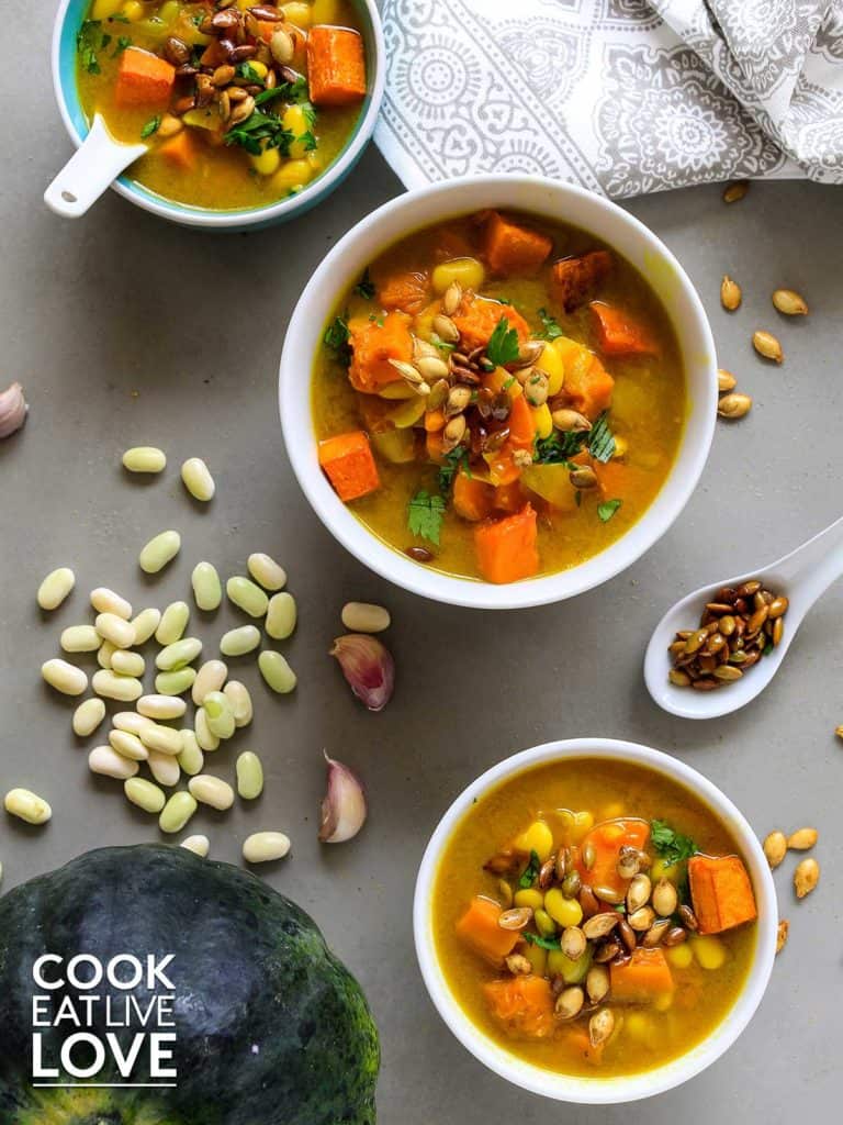 Three bowls of healthy pumpkin soup on gray background.  White beans and toasted pumpkin seeds around the bowls.