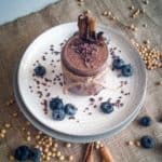 Jar of cacao chia pudding on white plate garnished with cacao nibs and blueberries.