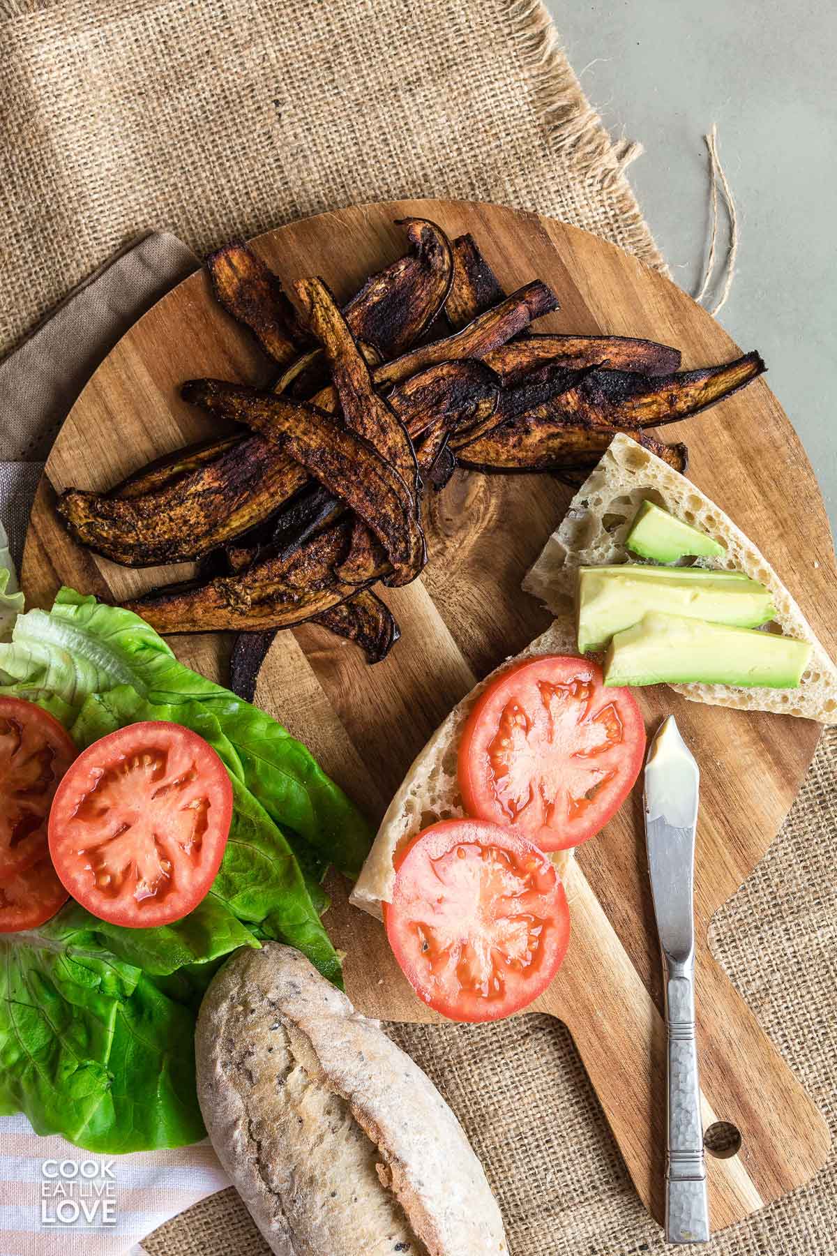 Slices of eggplant bacon on a cutting board with ingredients to make a sandwich.