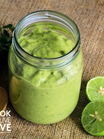 Jar on creamy avocado sauce with limes and parsley on burlap.