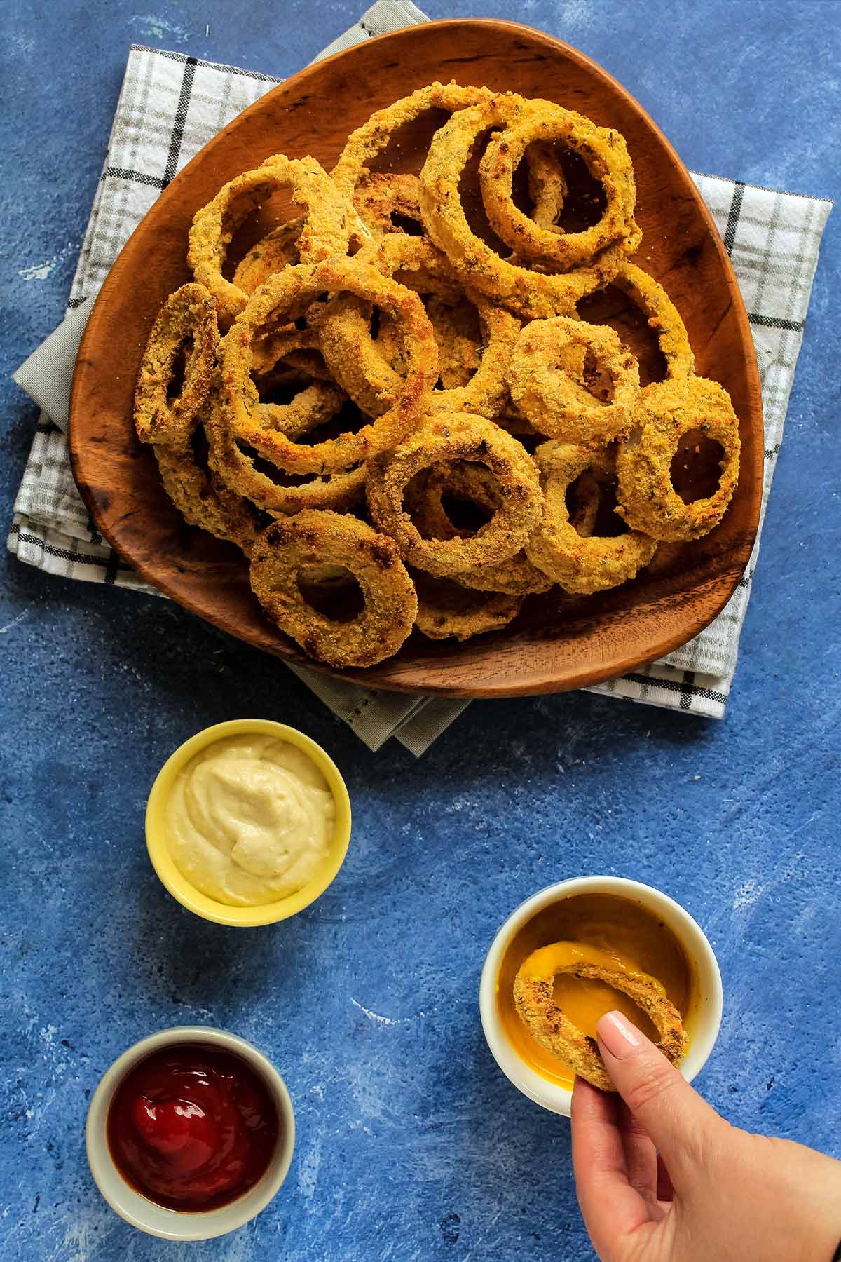 Platter of onion rings with three sauces on the table and a hand dipping one in sauce.