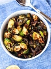 Cooked brussel sprouts in a white bowl on blue surface with spoon and lemon in frame.