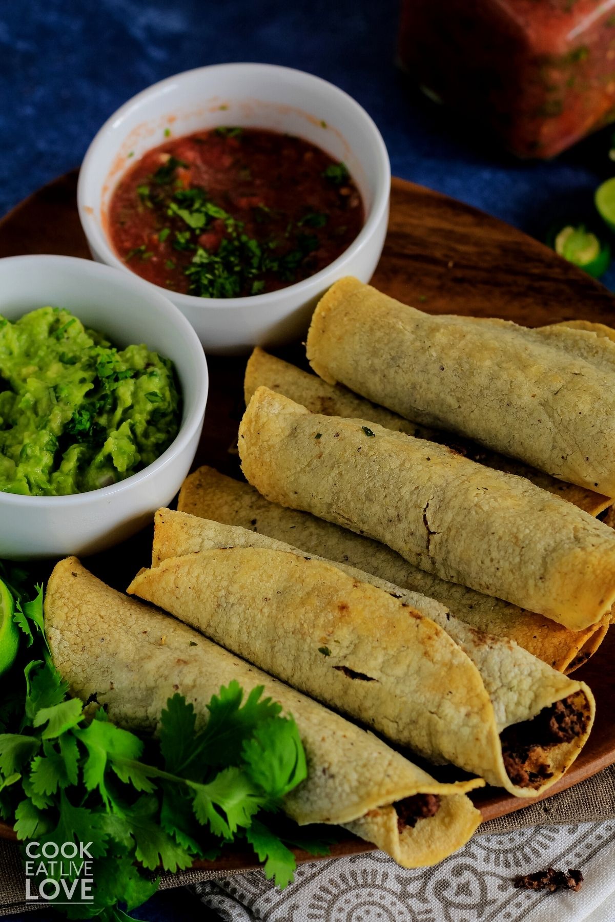 Vegan taquitos on a platter on the table with bowls of salsa and guacamole.