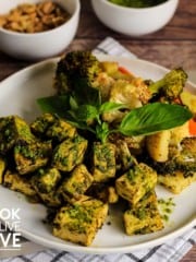 Overhead view of baked tofu and veggies on white plate with walnuts and pesto in small bowls behind the plate.