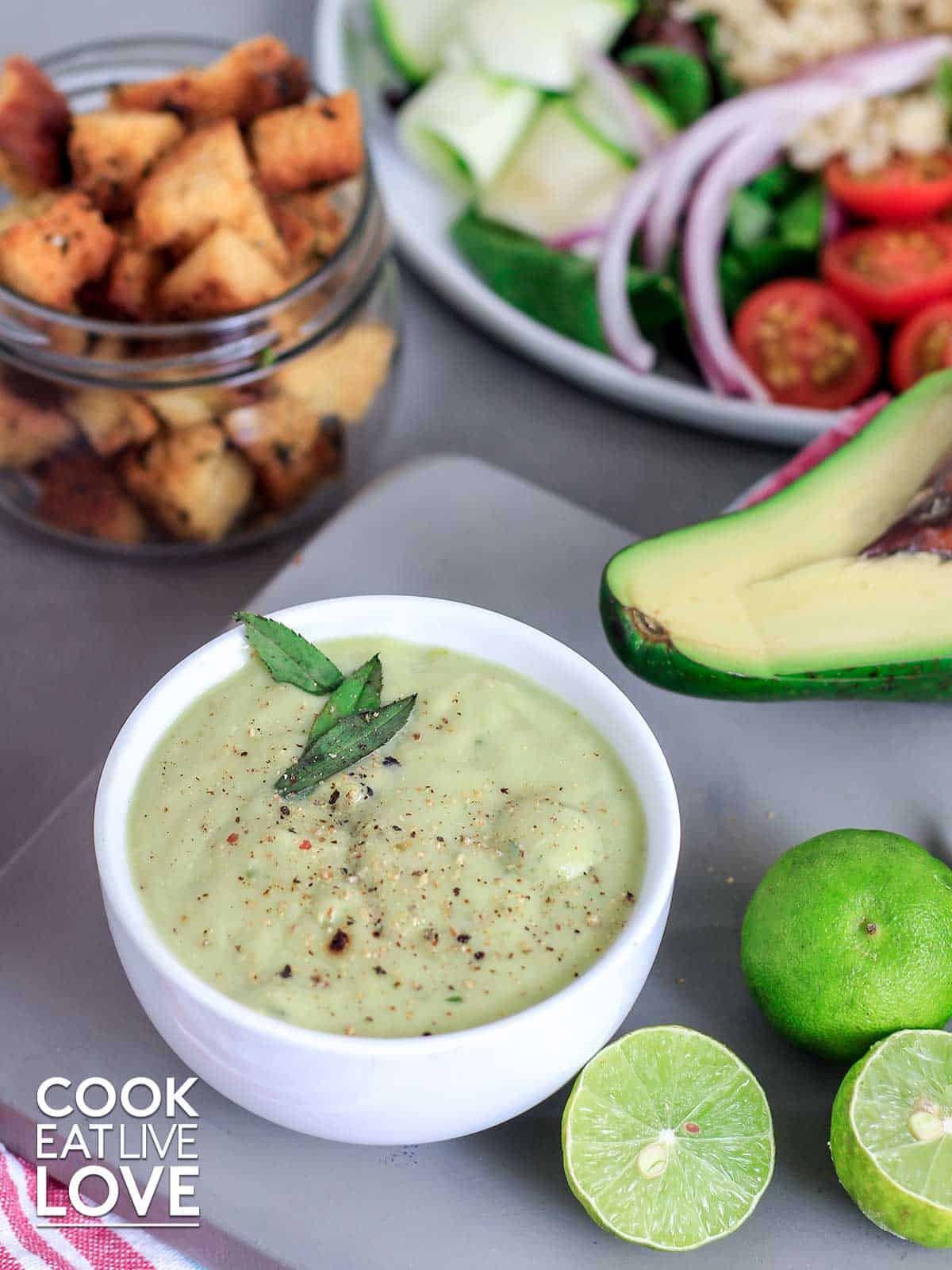 Avocado lime dressing served up in small white bowl with herb garnish and limes