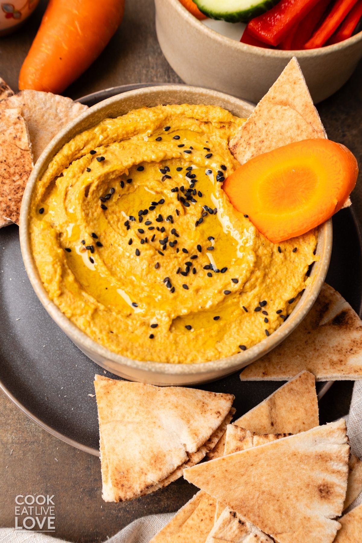 A bowl of carrot hummus topped with black sesame seeds a sliced carrot wedge digging in.