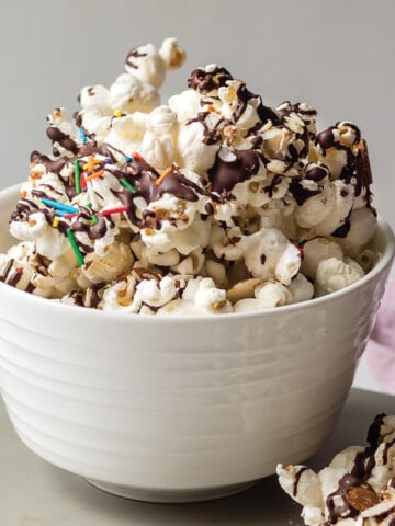 Chocolate drizzled popcorn in a small white bowl and some spilling on table in front.