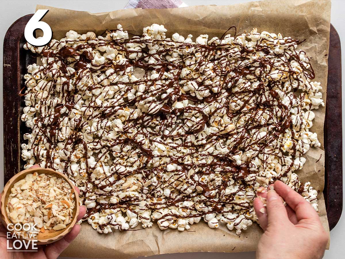Adding nuts to baking sheet of chocolate drizzled popcorn.