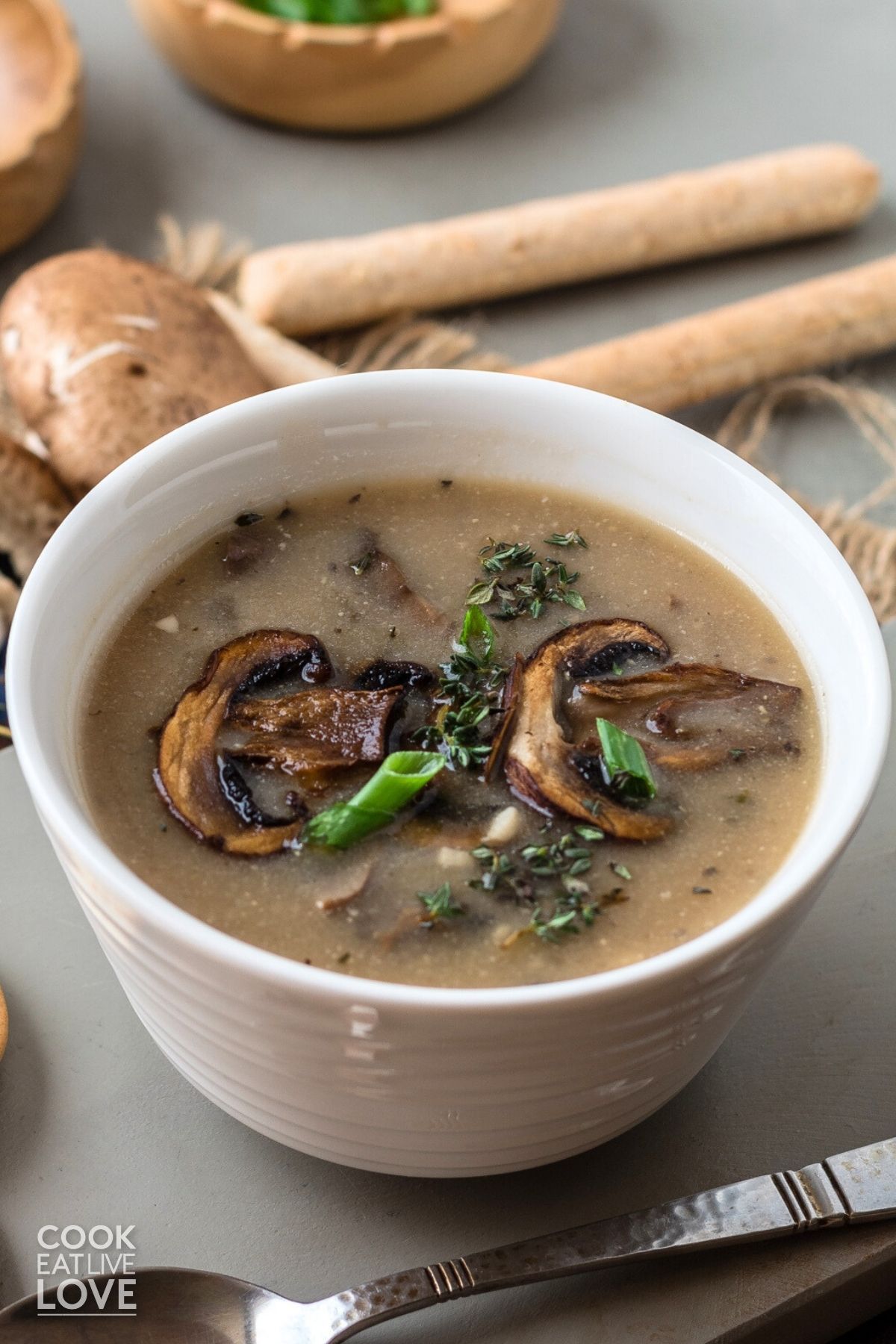 A bowl of mushroom soup without cream with breadsticks and whole mushrooms in the background. 