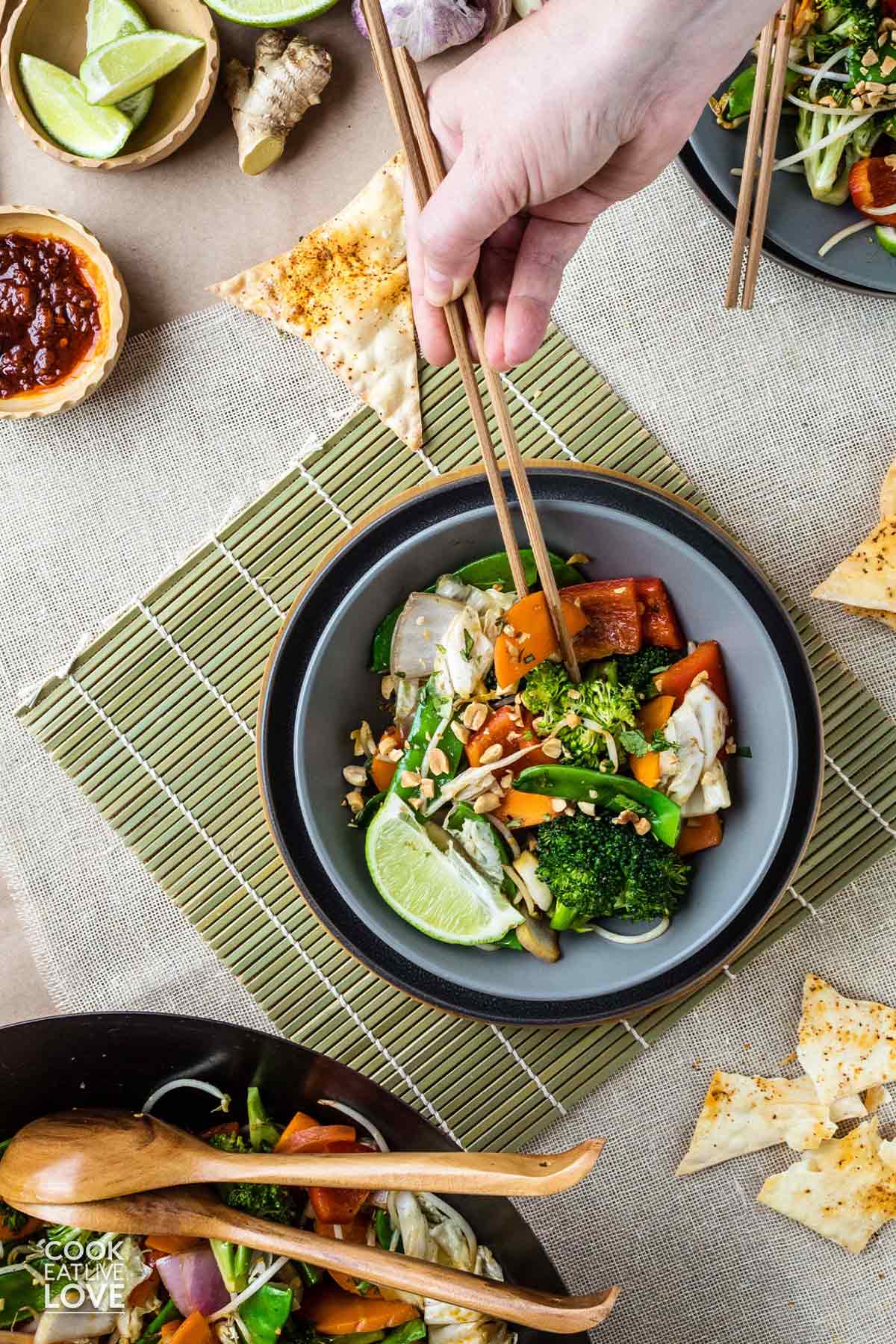 A bowl of spicy stir fry on the table with a hand holding chopsticks reaching in for a bite.