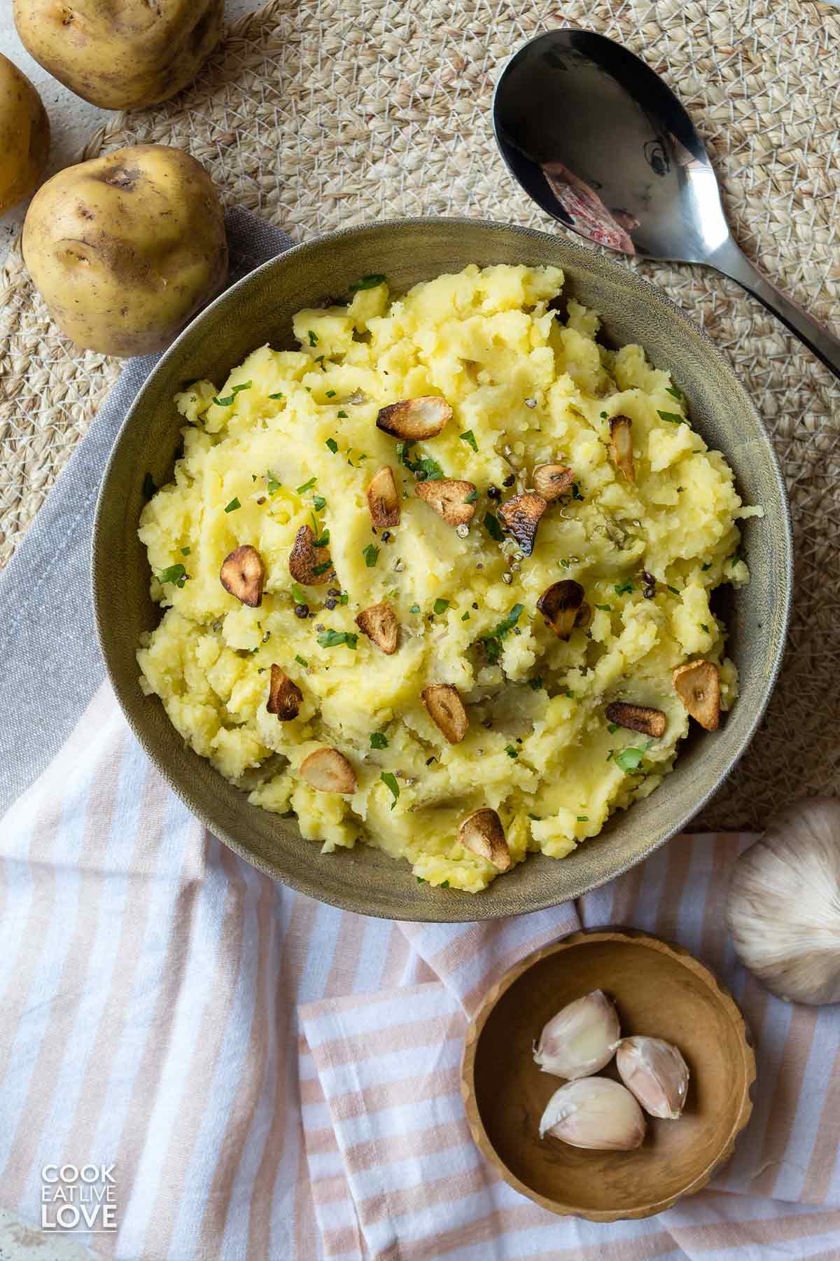 Mashed potatoes without butter served up on a table with whole potatoes and fresh garlic next to it.