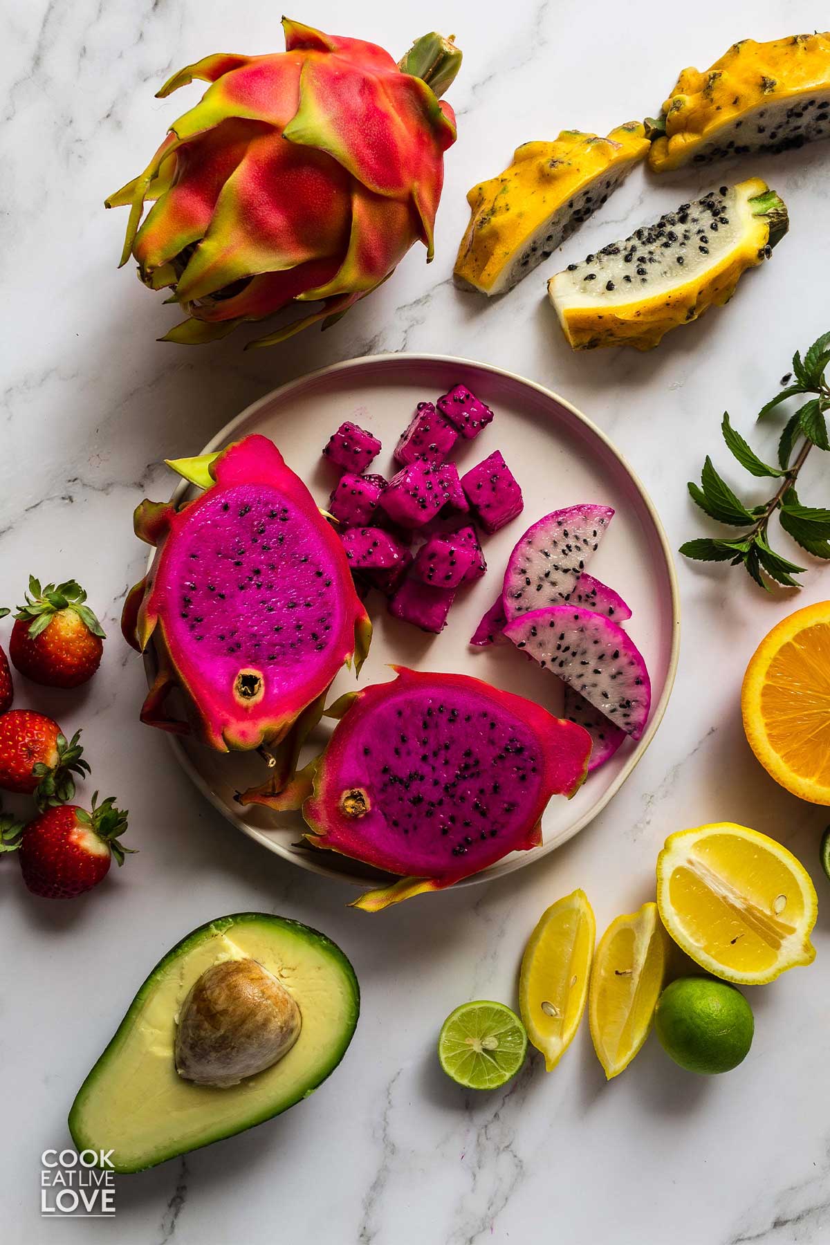 Overhead of dragon fruit whole and cut on the table surrounded by citrus, avocado, strawberries and other fruits.