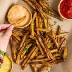 A hand dipping a baked French fry into a sauce.