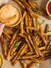 A hand dipping a baked French fry into a sauce.
