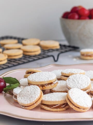 Peruvian alfajores cookies on a platter on the table with bowl of fruit and cookies on a cooling rack in the background.