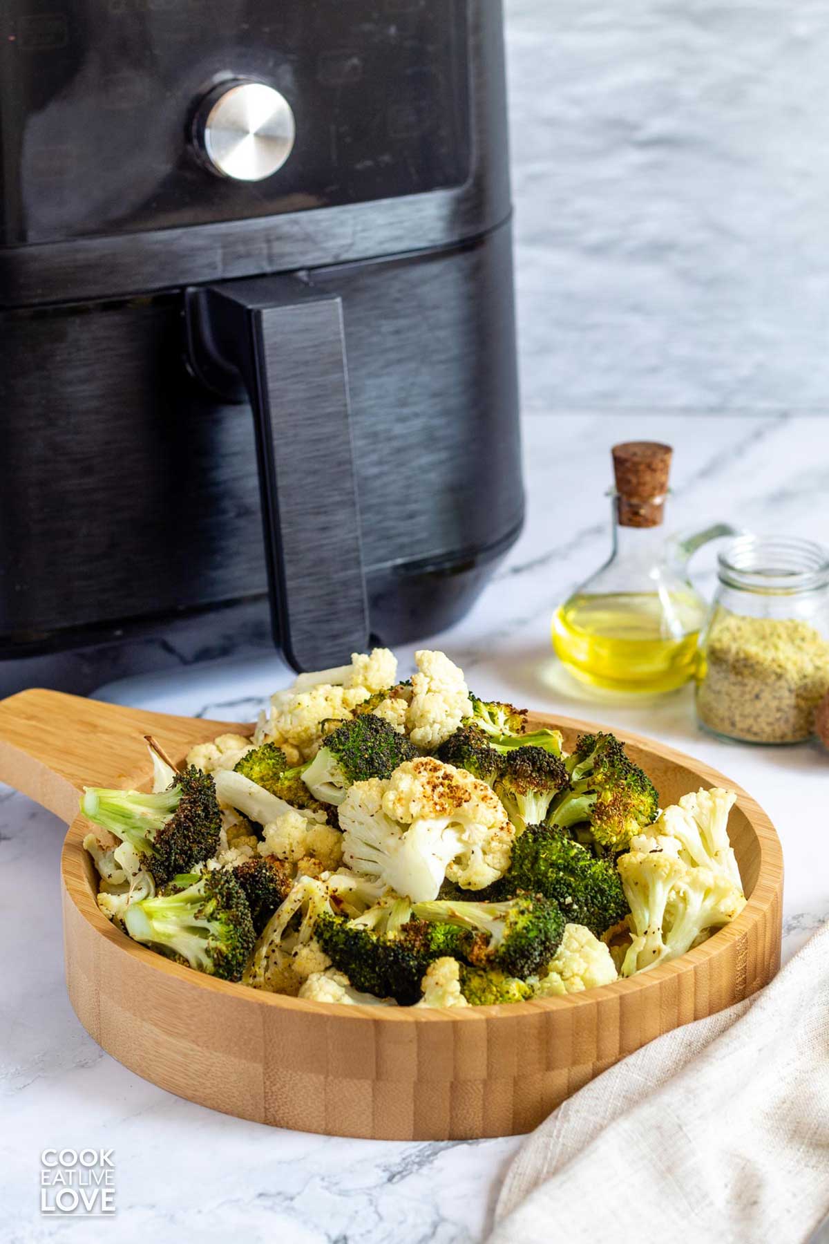 Broccoli and cauliflower served up in a wooden bowl in front of an air fryer.