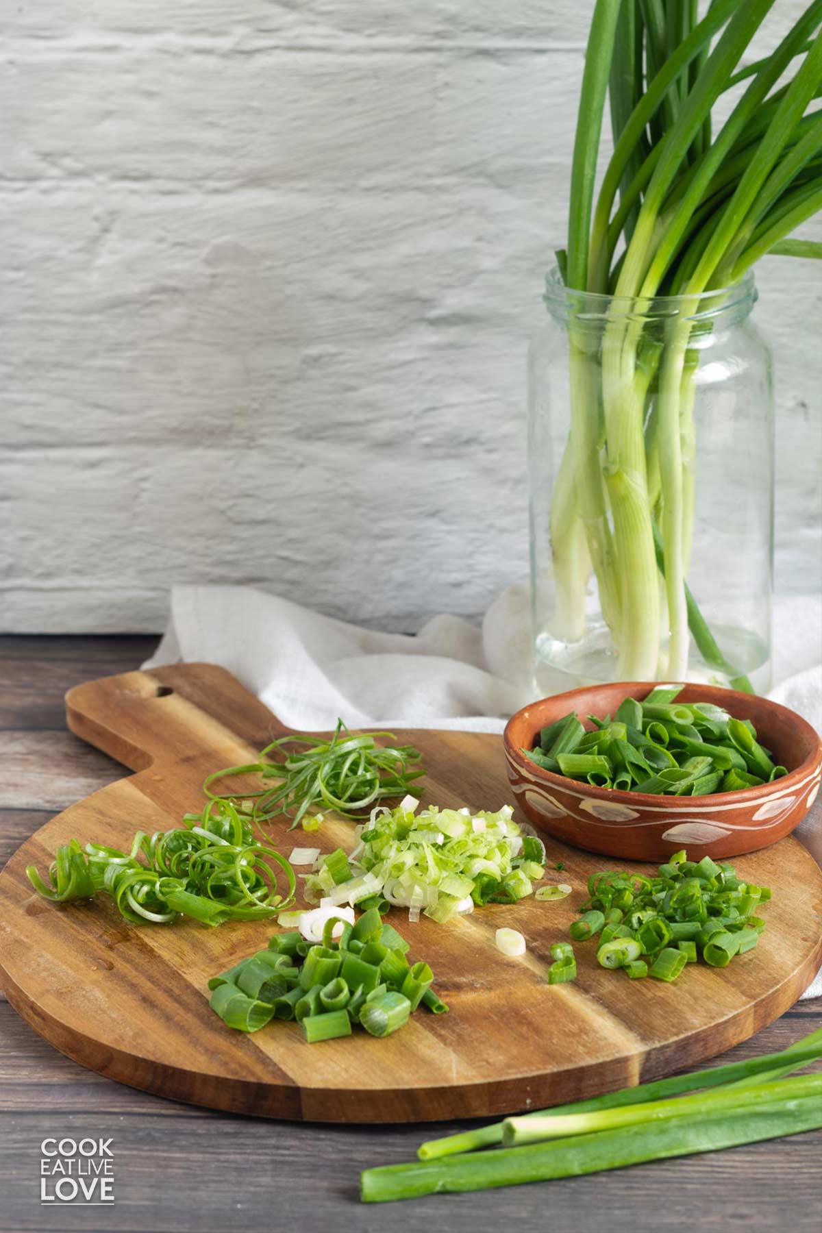 Different cuts of green onions on a round cutting board with green onions in a jar in the background.