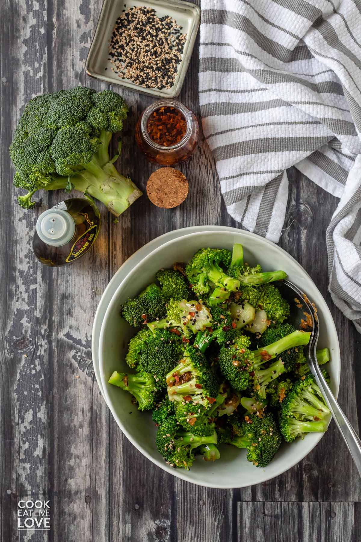 Asian broccoli on the table in a bowl with a bowl of sesame seeds, sesame oil, and a jar of chili oil