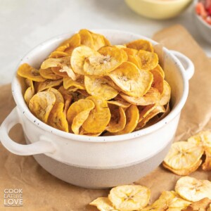 Baked plantain chips in a small bowl with more on parchment paper in the front.