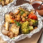 Baked vegetables in foil on the table with a small bowl of sauce in the background.