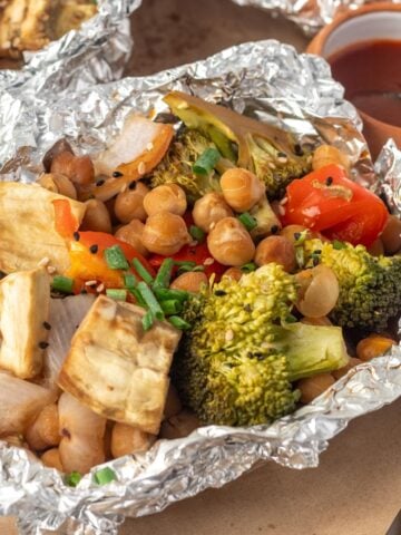Baked vegetables in foil on the table with a small bowl of sauce in the background.