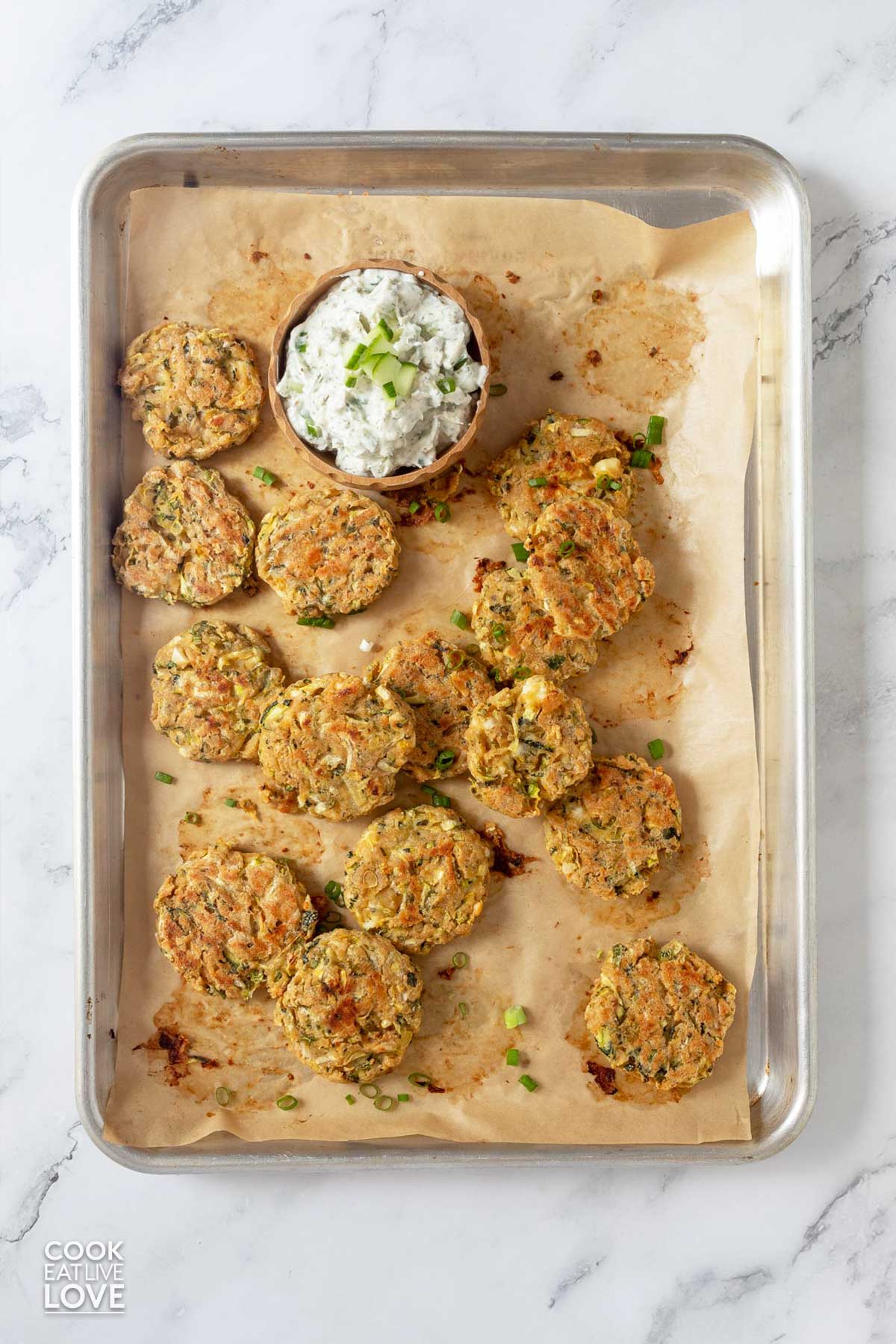 Cooked zucchini fritters on a baking pan with a bowl of yogurt dip on it for dipping.