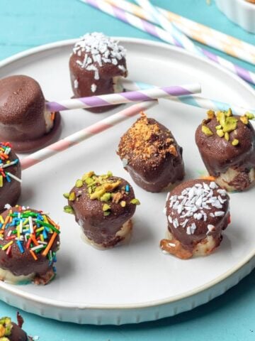 A plate of frozen banana bites on the table dipped in chocolate and topped with assorted sprinkles.