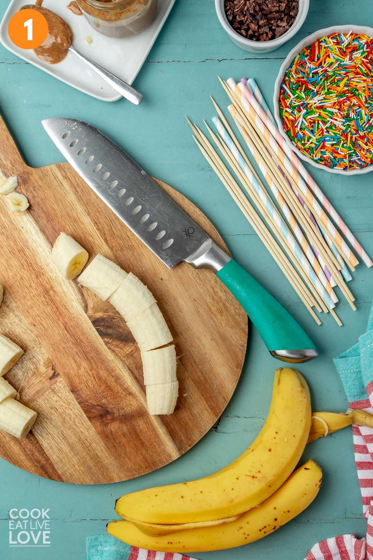 Sliced bananas on a round cutting board with toppings, banana peel, and a knife to the side.