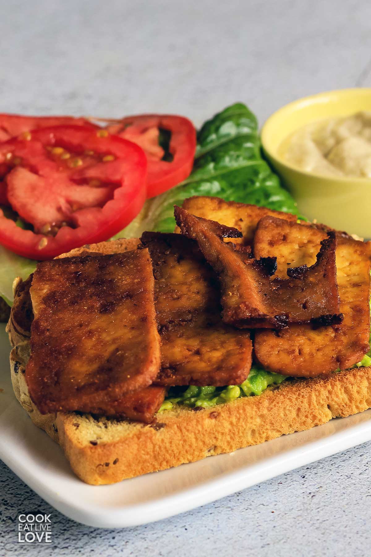 A piece of bread topped with marinated tofu with another slice of bread in the back with lettuce and tomato.