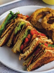 Tofu blt sandwich served up on plate with onion rings and creamy white sauce.