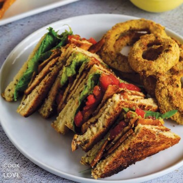 Tofu blt sandwich served up on plate with onion rings and creamy white sauce.
